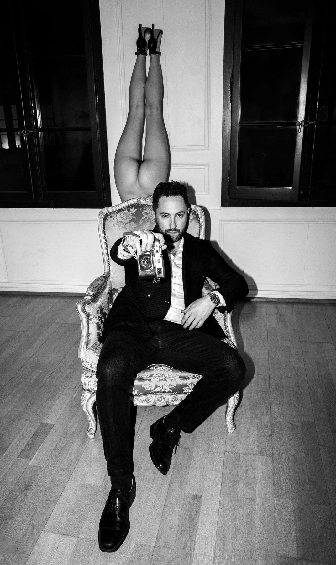 Jeremy Spierer seated in an armchair with his camera, a model's legs raised behind him